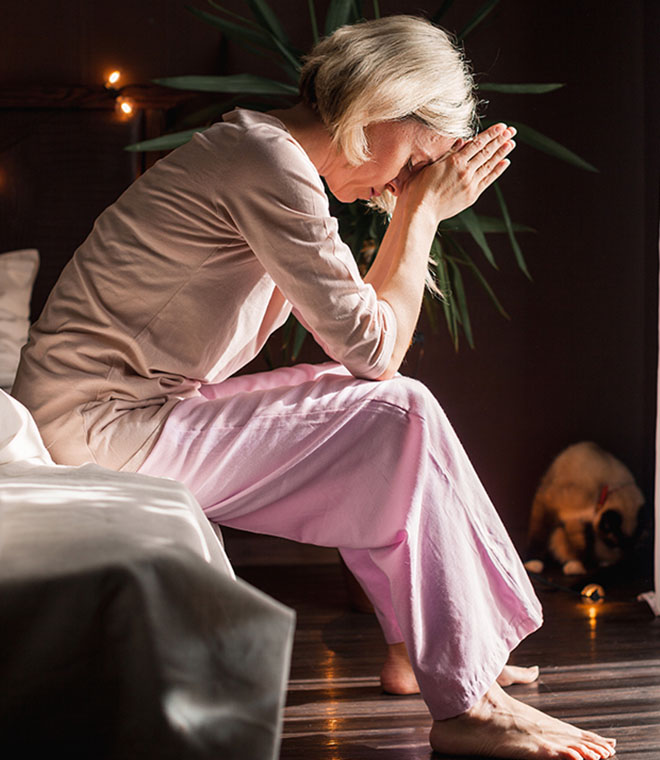 Older white woman sitting on bed holding her head