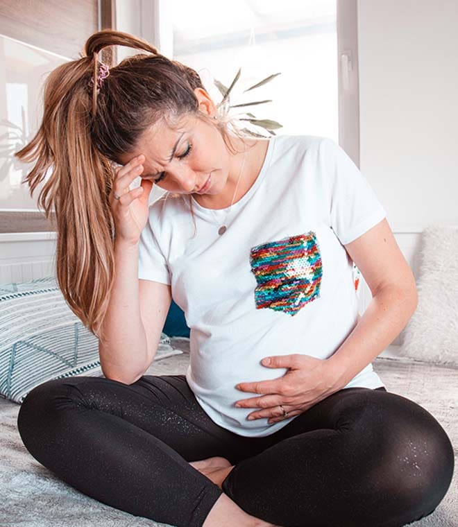 Pregnant white woman sitting on bed holding her belly and her head
