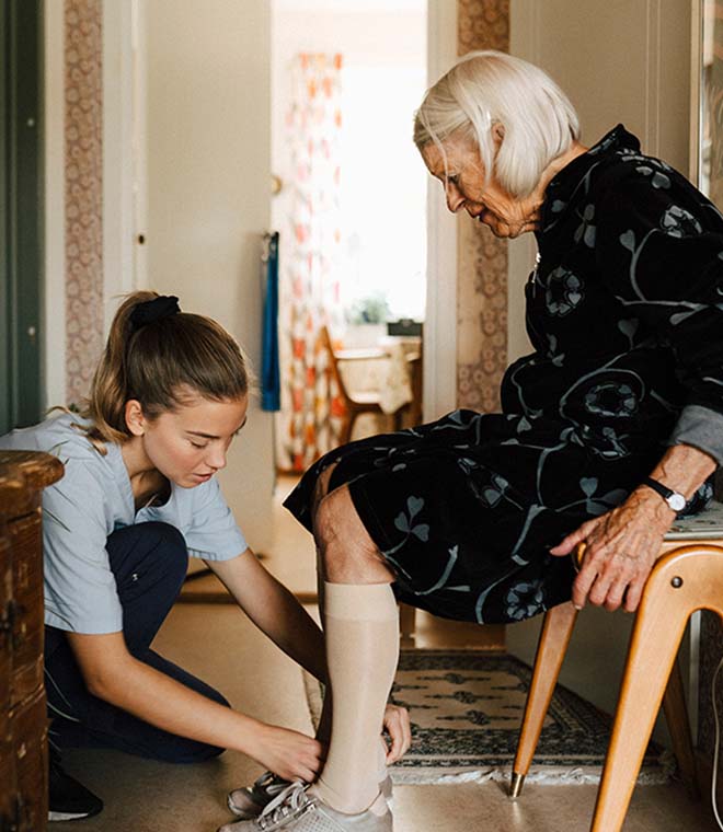 Female caregiver helping senior woman put on a shoe at home