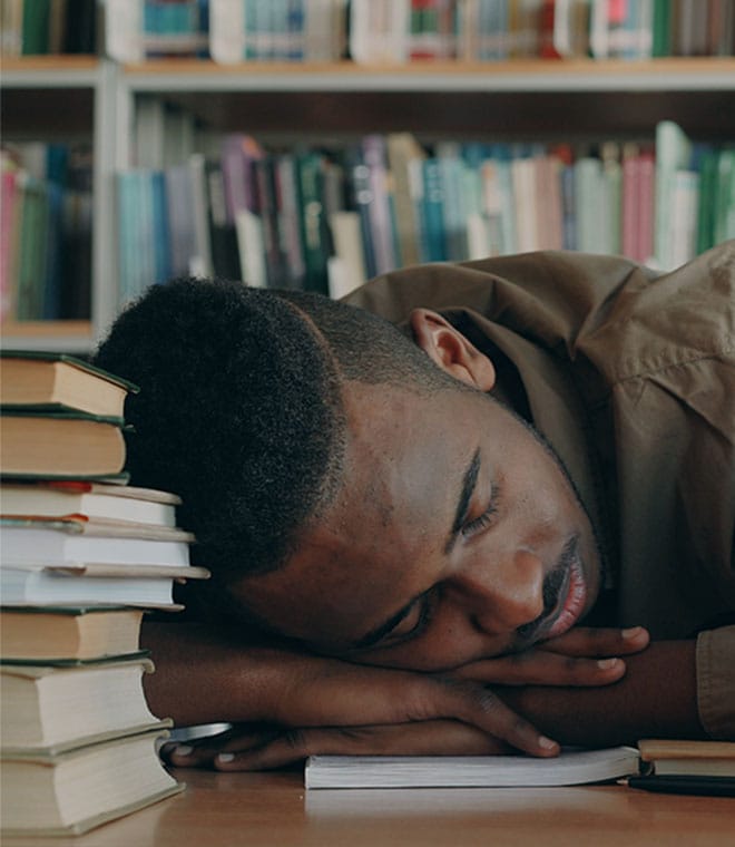 Black man resting his head on a desk full of books