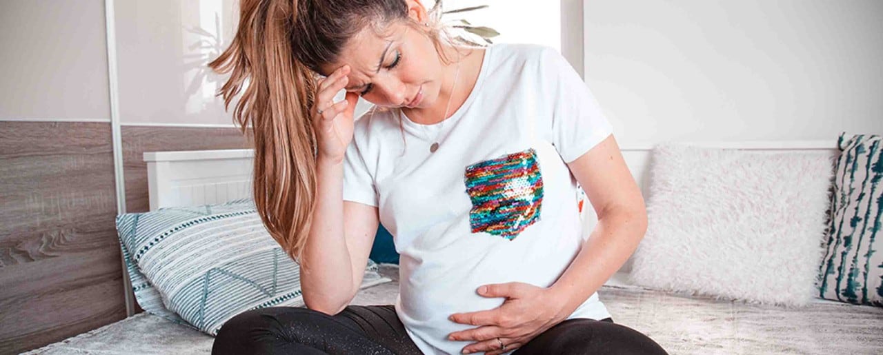Pregnant white woman sitting on bed holding her belly and her head